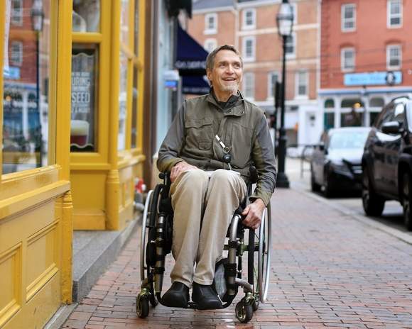 Exterior daytime photo of a man in a wheelchair on a cobblestone sidewalk near a yellow building entrance. The man is middle aged, Caucasian with short light brown greyish hair, and is dressed in neutral colors. He is looking away from the camera and smiling.