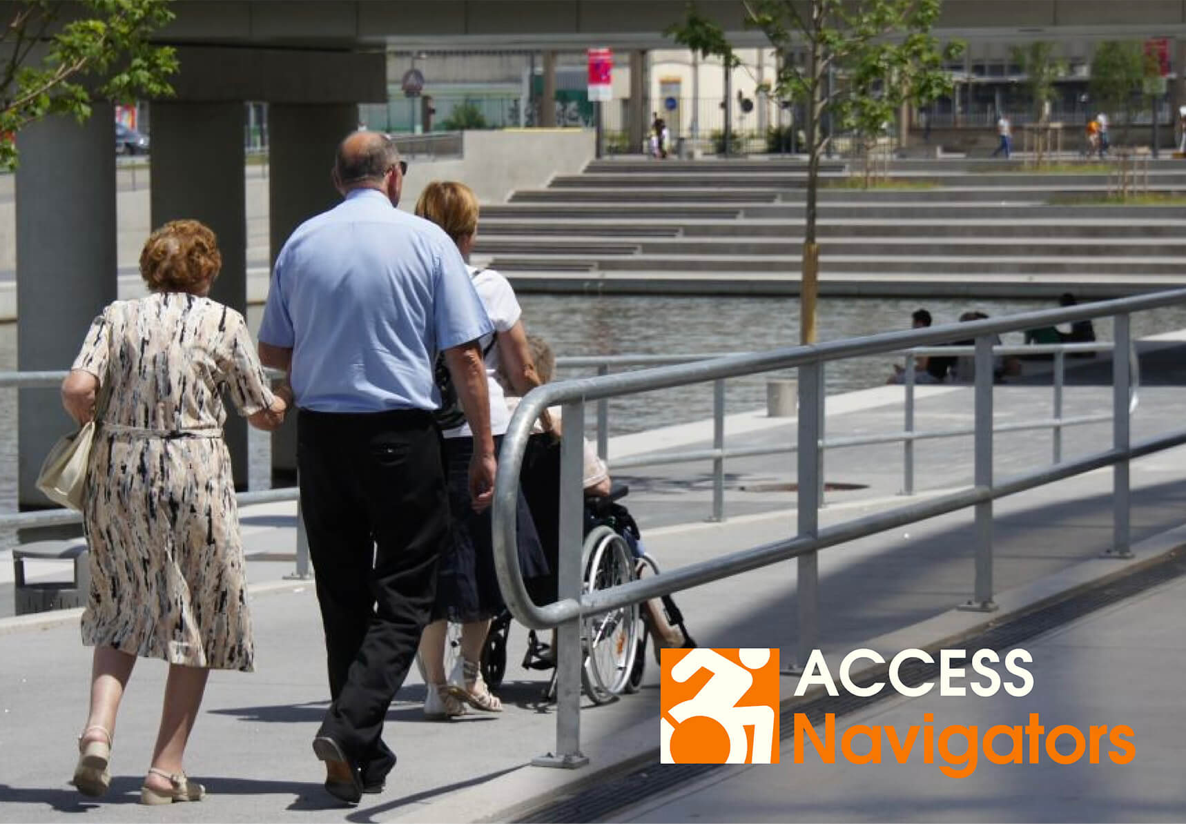 Taken from behind the subjects, an elderly man and woman follow behind someone pushing a person seated in a wheelchair along a stretch of concrete walkway with guardrails on either side.