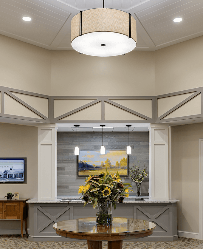 Interior view of an entrance area to a senior living facility. A high ceiling frames a neutral colored room with beige and grey trim, with a grey wood reception area desk featured behind a dark wood circular small table with a vase of sunflowers.