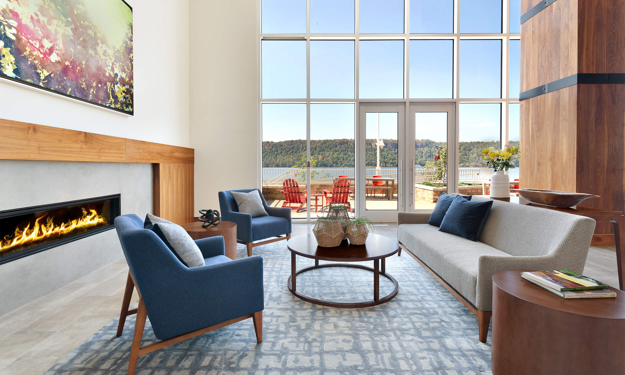 Interior view of a community room in a luxury apartment complex. The room is bright and sunny with natural light coming from large windows, and a modern fireplace on the left is flanked by blue midcentury modern upholstered chairs.