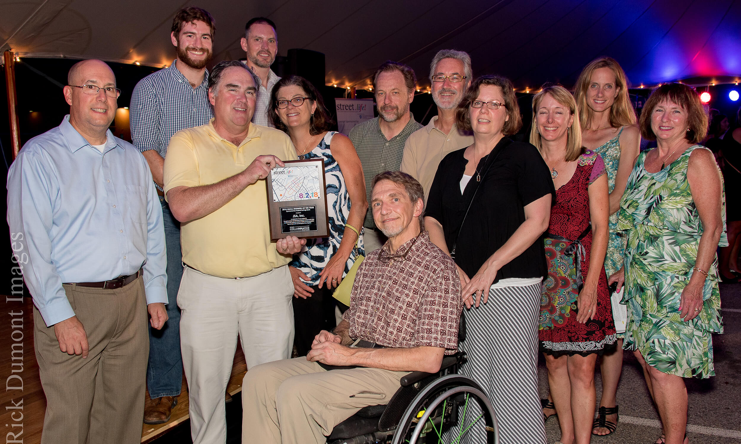 A group of twelve people are gathered around and looking into the camera while smiling, as they've won an award. The group is a mix of men and women, with a man in a wheelchair in the center of the group. The man in a yellow shirt to the left of him is holding an awards plaque.