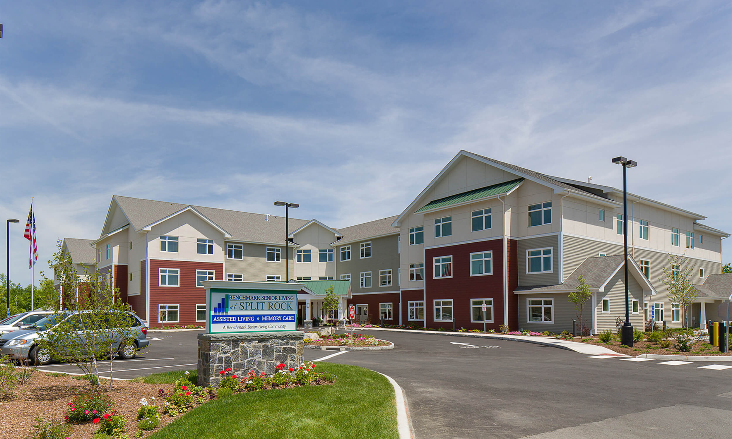 Exterior view of the main entrance to Benchmark Village at Split Rock senior living facility. The multi-story building features a red and white facade with a drive-thru parking lot area in front.