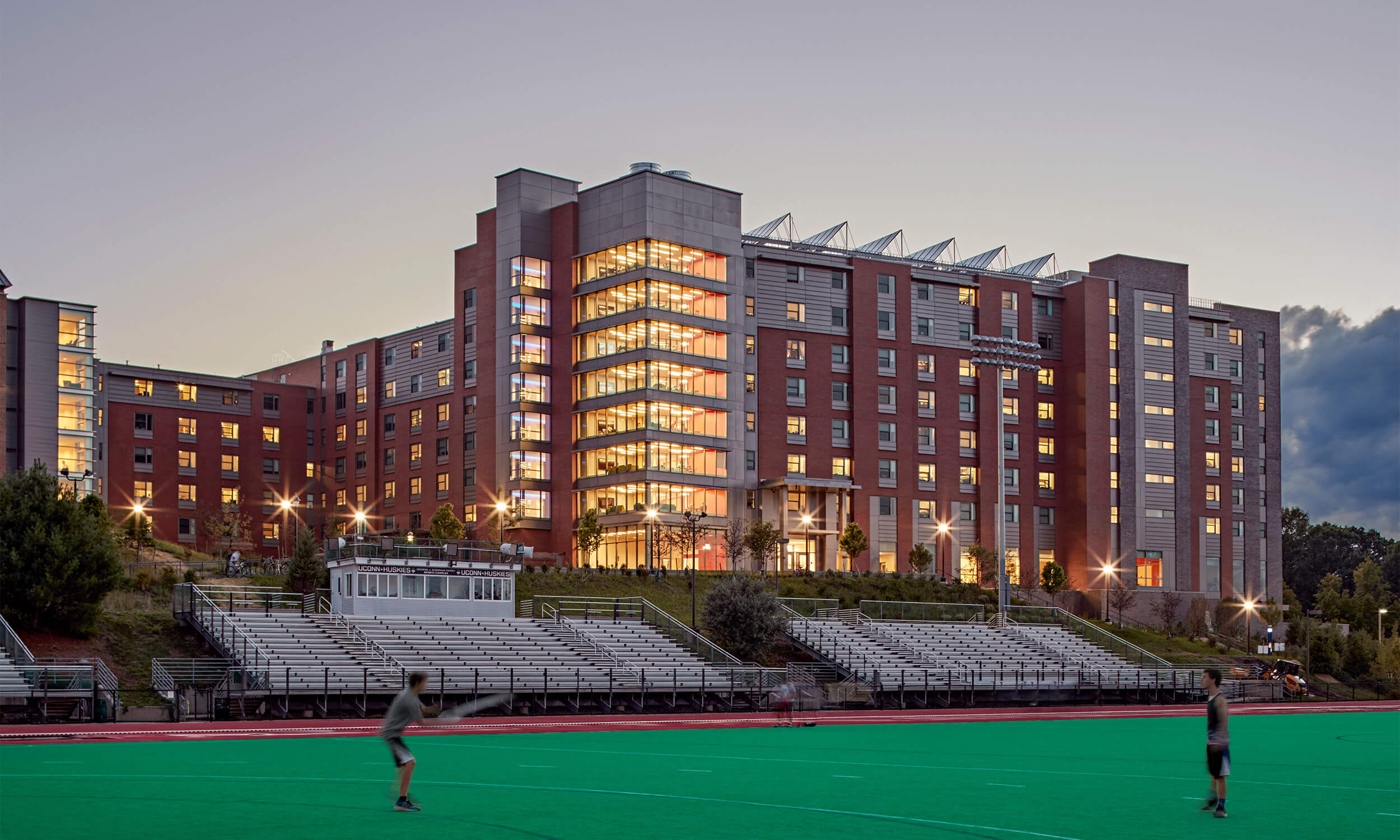 Exterior view of a college campus building as seen across an athletic field. The photo appears to be taken at dusk, with lighting illuminating the building from within.
