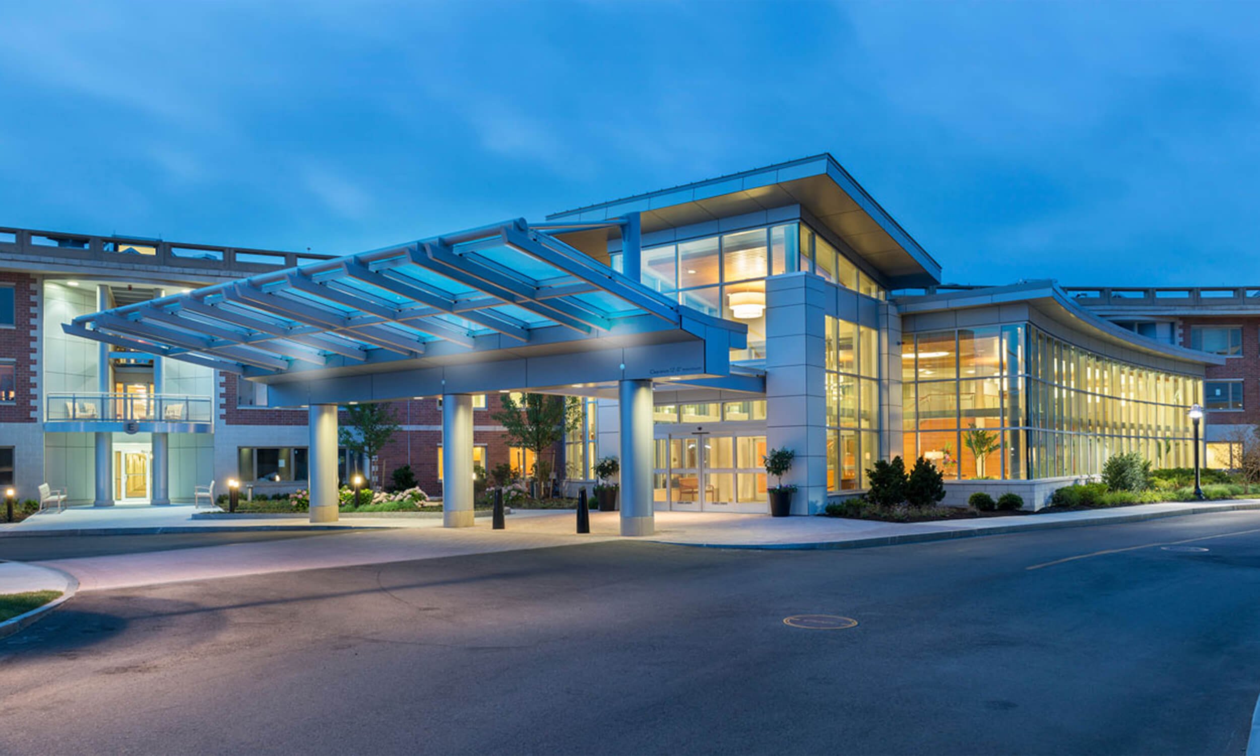 Exterior view of the entryway to a senior living community. The photo is taken at dusk, with lights inside the building illuminating it from within. Large floor-to-ceiling glass walls are featured on the facade.
