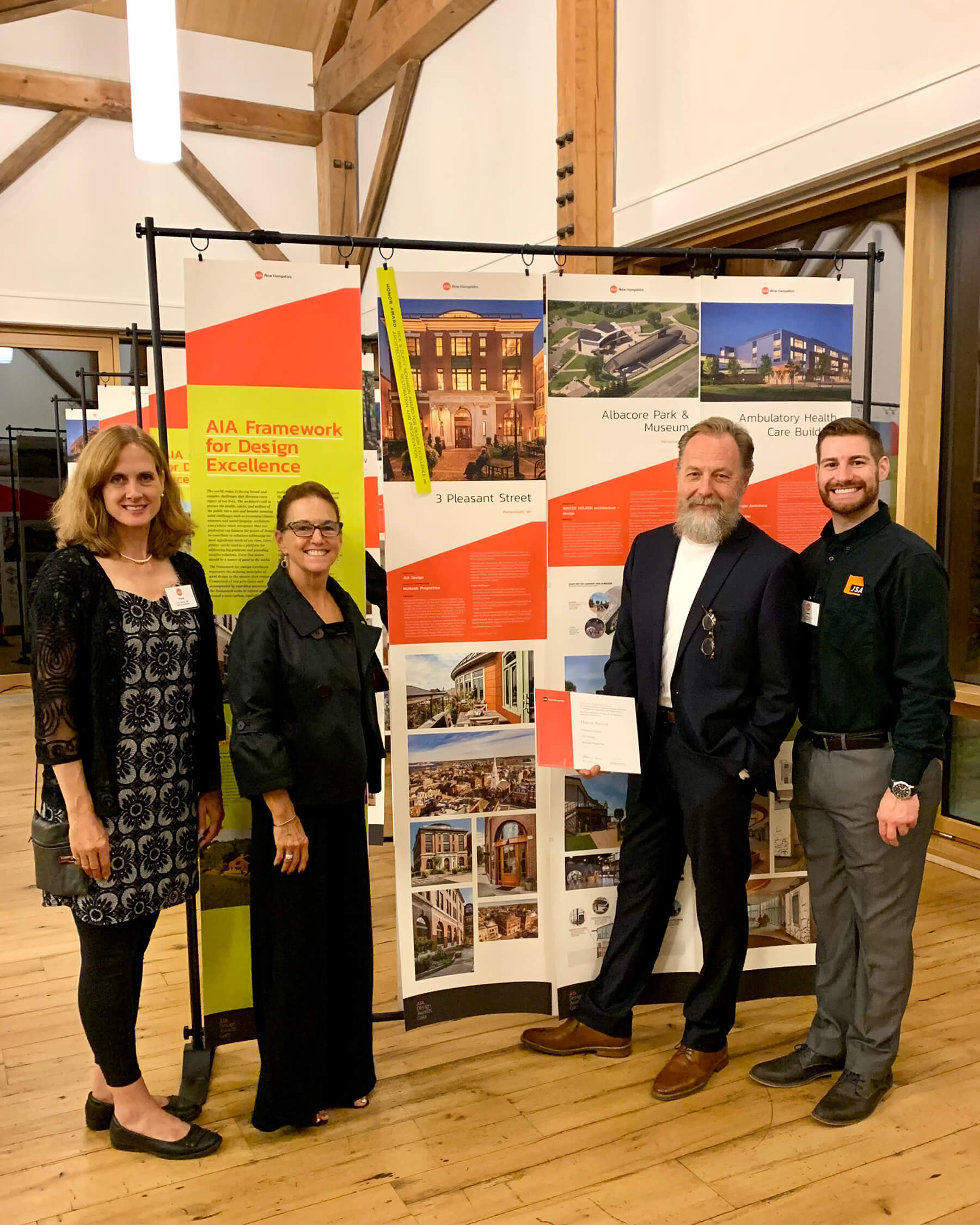 A group of four people (two men and two women) stand next to a presentation exhibition board. They are at an architecture awards event.