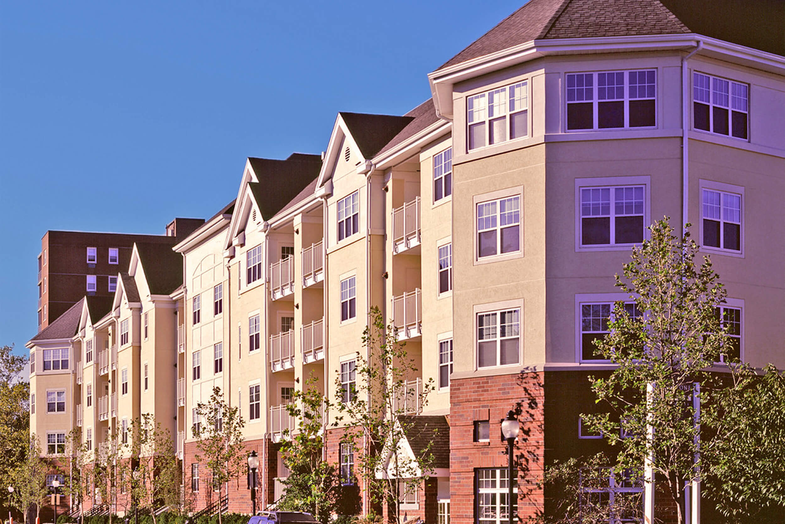 Exterior daytime street view of a multi-unit housing complex. The facade of the building features neutral colored siding with brick accents.