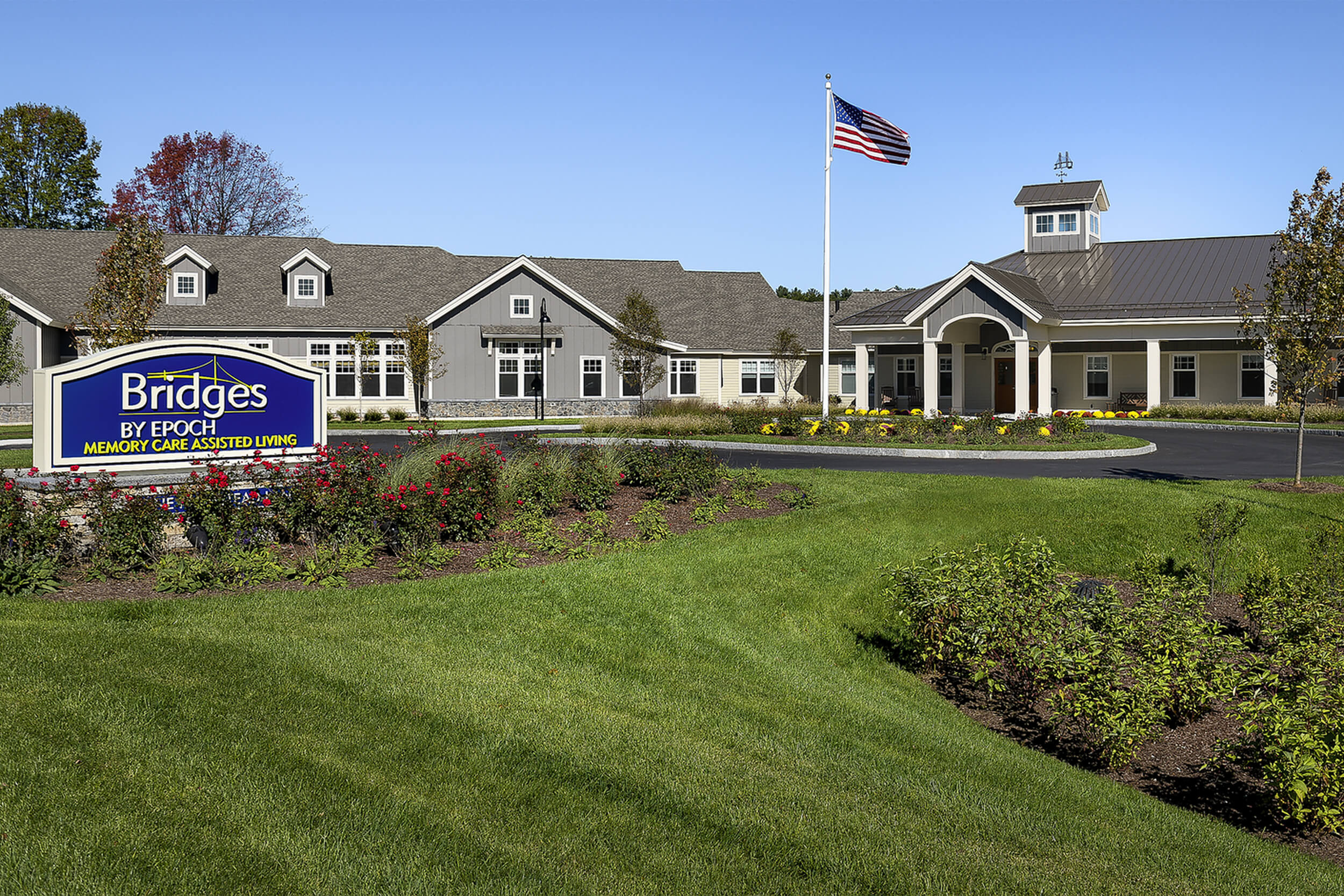 Exterior daytime view of a senior living facility. A large grassy lawn stretches in front of the building with a sign reading "Bridge by Epoch"