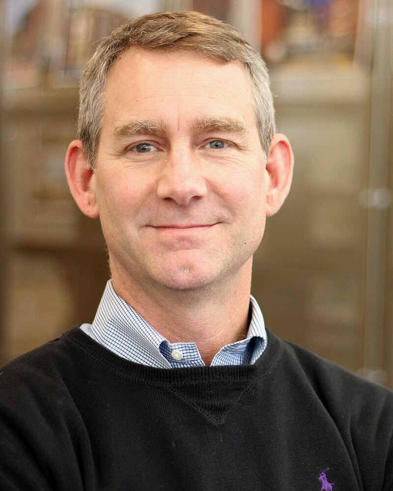 Headshot from the shoulders up of a middle aged Caucasian man. He is looking into the camera and smiling with his mouth closed. He has light sandy blonde hair cut short, and is wearing a collared light blue shirt under a navy sweater.