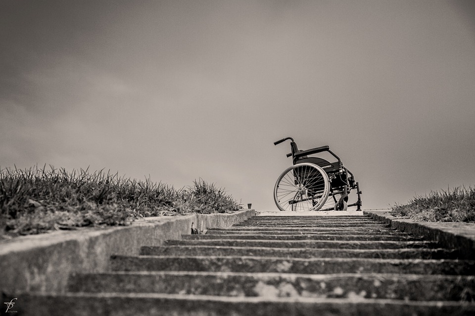 Black and white photo of a lone wheelchair at the top of a set of shallow concrete steps outdoors.