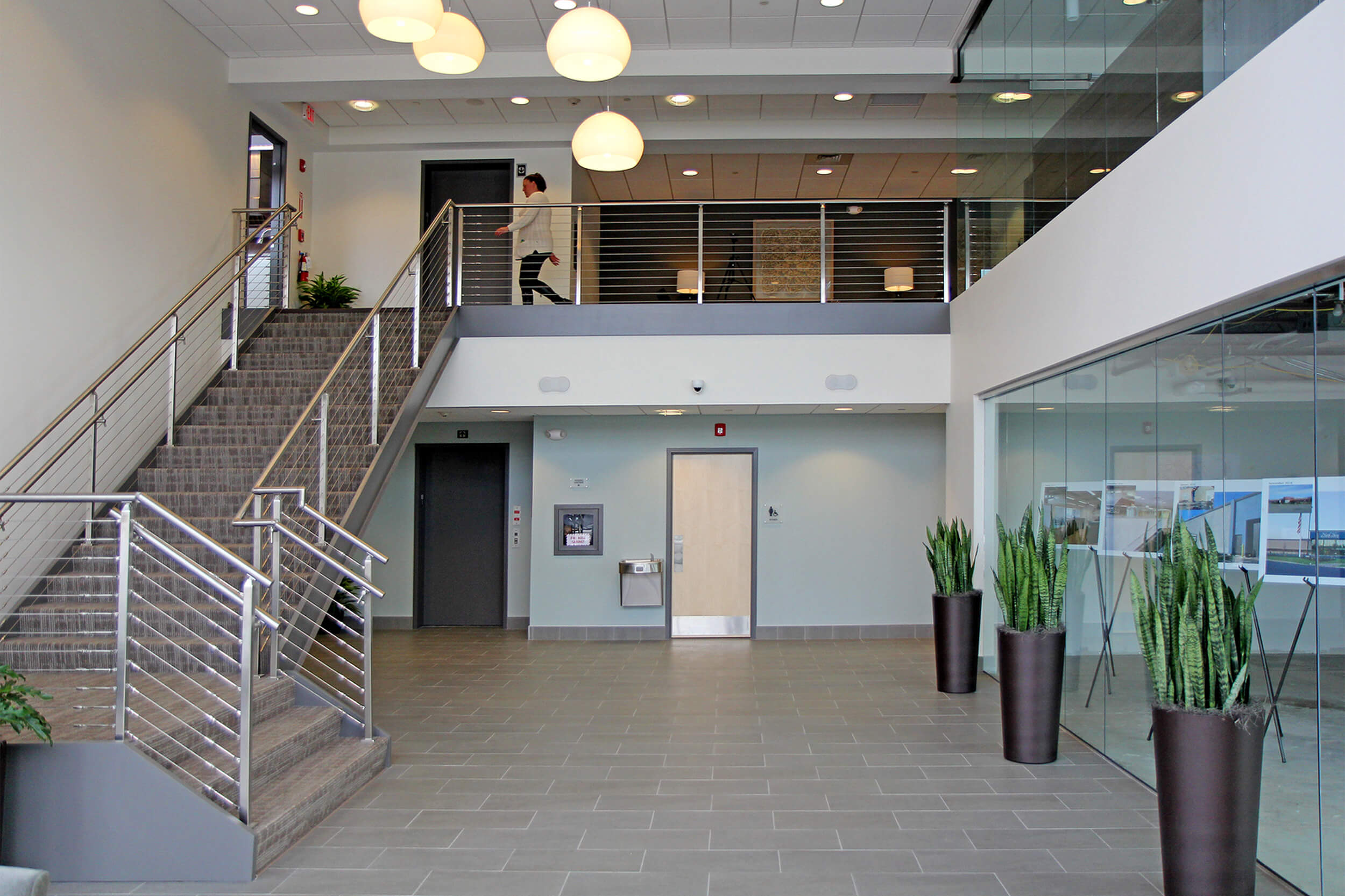 Interior photo of the lobby entrance to a coporate office space. The floor and walls are neutral grey tones, and a staircase on the left leads to a second floor. Plantings and a reception desk can be seen on the right side of the lower level.