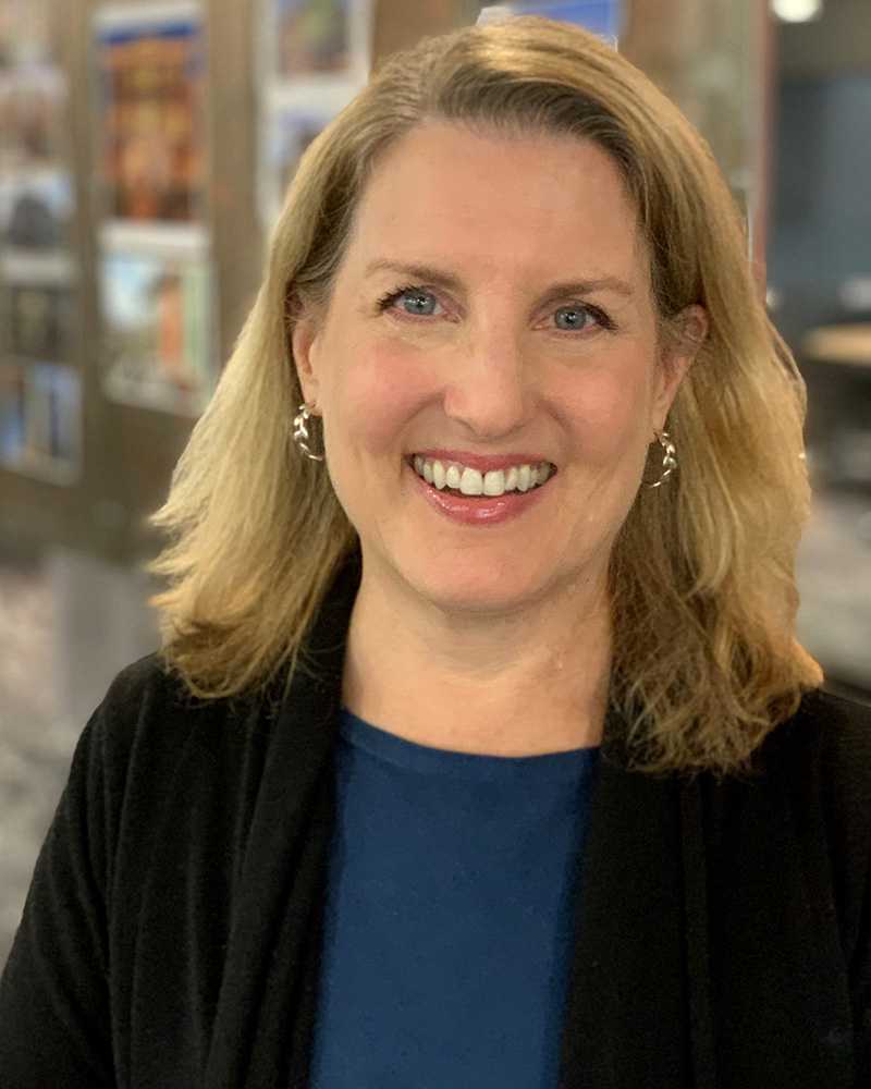 Headshot photo from the shoulders up of a middle aged Caucasian woman. she has shoulder length straight blonde hair and is smiling into the camera. She wears a blue shirt with a black cardigan.