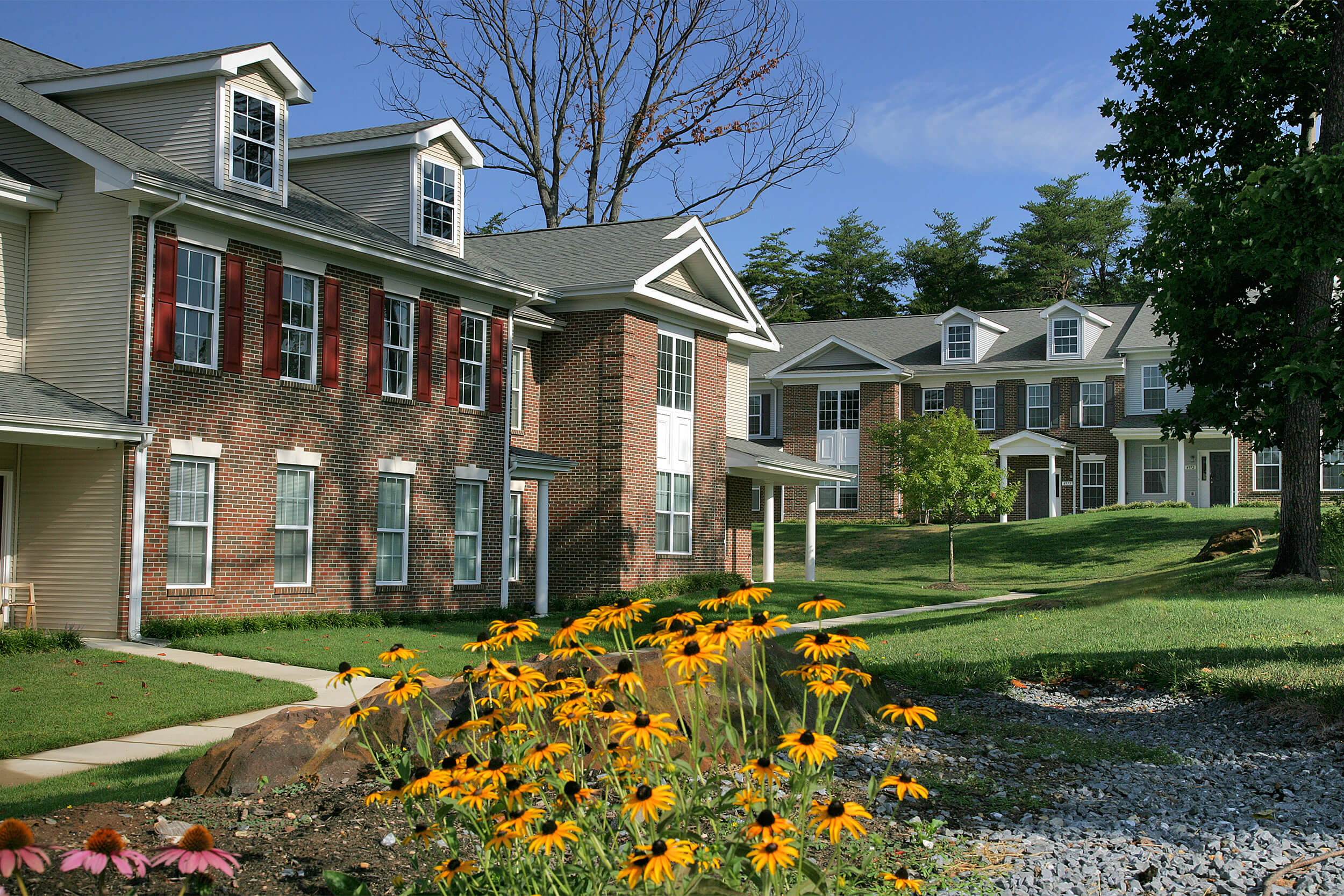 Exterior daytime view of multi-family housing units. The facade of the buildings are brick with neutral siding and stone accents, and yellow flowers are seen in the foreground.