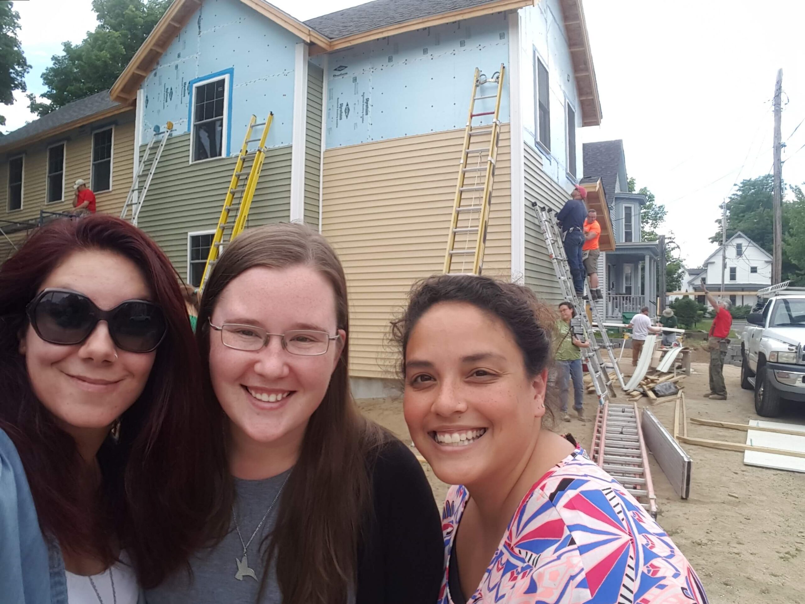 Three young women are gathered close together in front of a home under construction. They are looking into the camera and smiling.