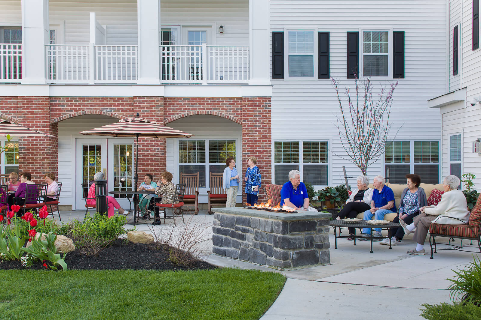 Outside view of a courtyard at a senior living facility. Several seniors are gathered in the courtyard at various picnic tables and lounge seating, socializing with one another.