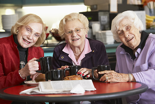 Three senior ladies are gathered around a table enjoying coffee. They are looking directly into the camera and smiling. All three women are caucasian, have shorter haircuts in shades of grey/white, and are wearing sweaters.
