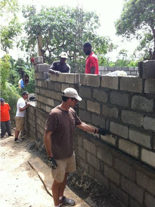 Photo showing several men building a concrete brick wall. The men are mostly black Haitian, with a Caucasian man wearing a baseball hat in the foreground smoothing out mortar among the bricks that are being laid.
