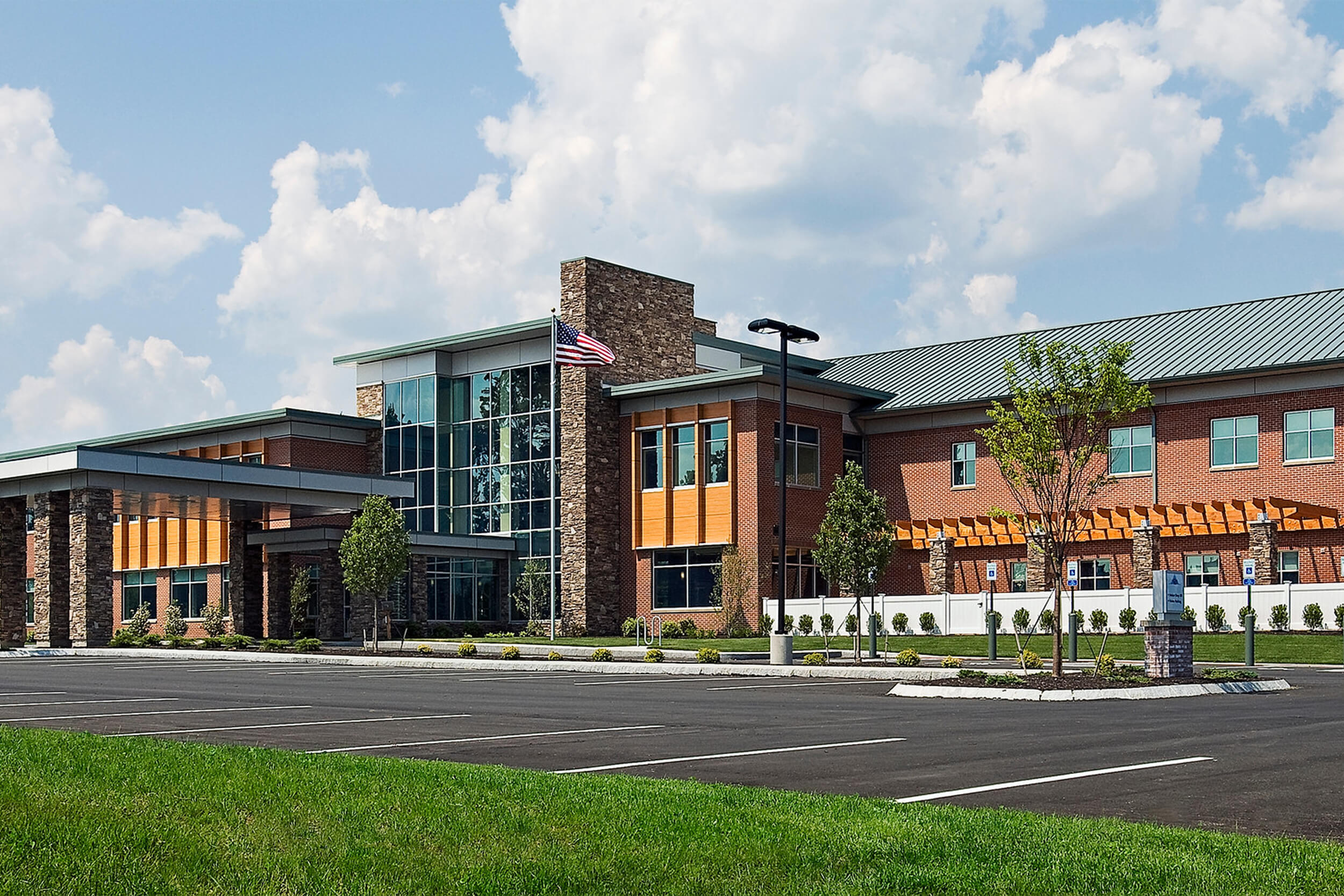 Exterior daytime view of a rehabilitation hospital facility. The building facade features dark brown siding with stone accents and large glass windows/walls.