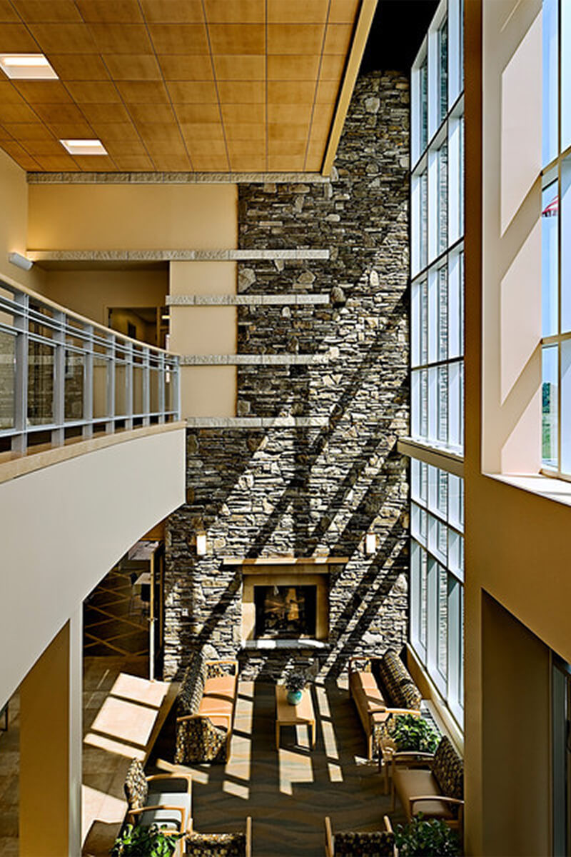 Interior photo of a lobby atrium area at a rehabilitation hospital facility. A large stone accent wall is next to floor-to-ceiling glass windows, letting in lots of natural light.
