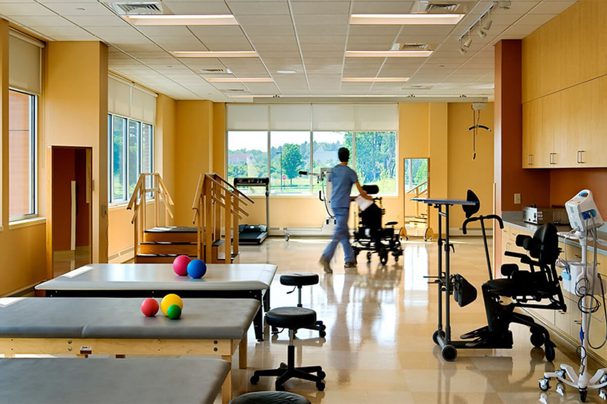 Interior view of a therapy room at a rehabilitation hospital facility. A staff member with their back towards the camera is pushing a person in a wheelchair past rows of physical therapy equipment.