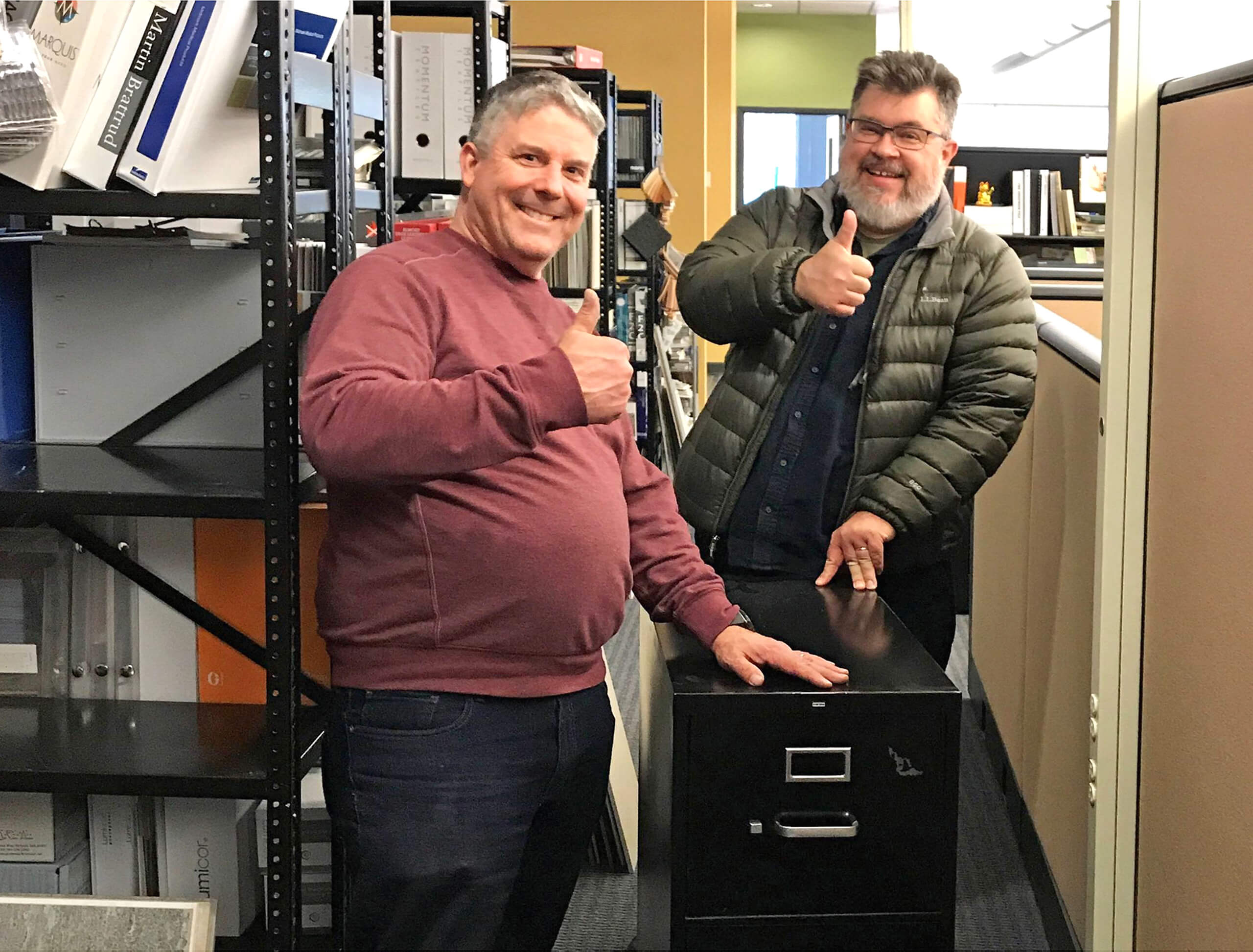 Two men are smiling and giving the thumbs up sign as they are touching a small black filing cabinet in an office environment.