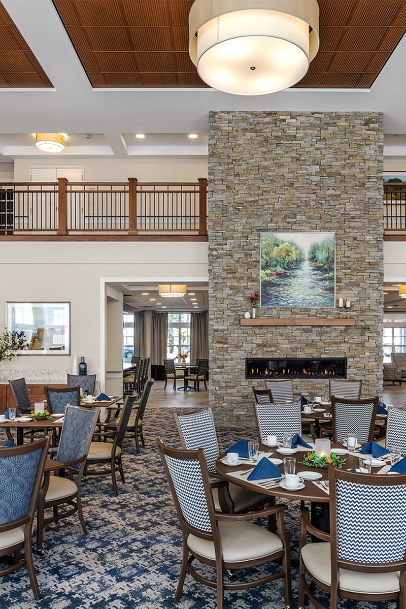 Interior photo showing a dining room area at a senior living facility. A large stone fireplace column is in the center of the room, with light blue-grey upholstered wood dining chairs around rectangular tables with place settings.