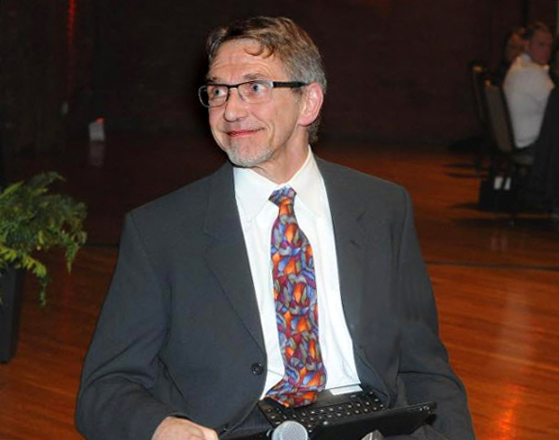 Photo of a man in a dark grey suit with white shirt and red pattern tie shot from the waist up. He is sitting in a wheelchair and looking away from the camera and smiling.