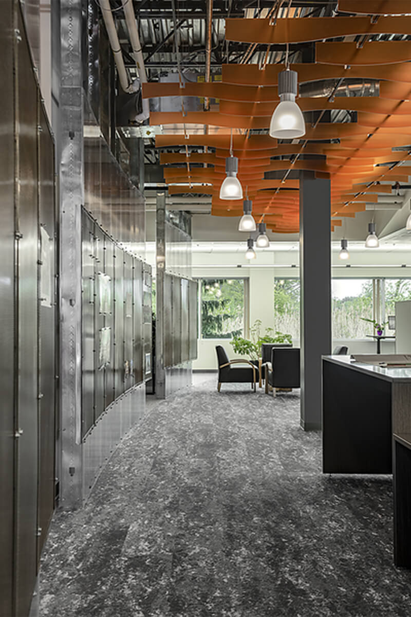 Interior view of an office space corridor flanking an open-layout area of working desks. The floor is a grey pattern carpet and orange ceiling accent panels bring a pop of color to the space. The left side features a communal work area pin-up wall.