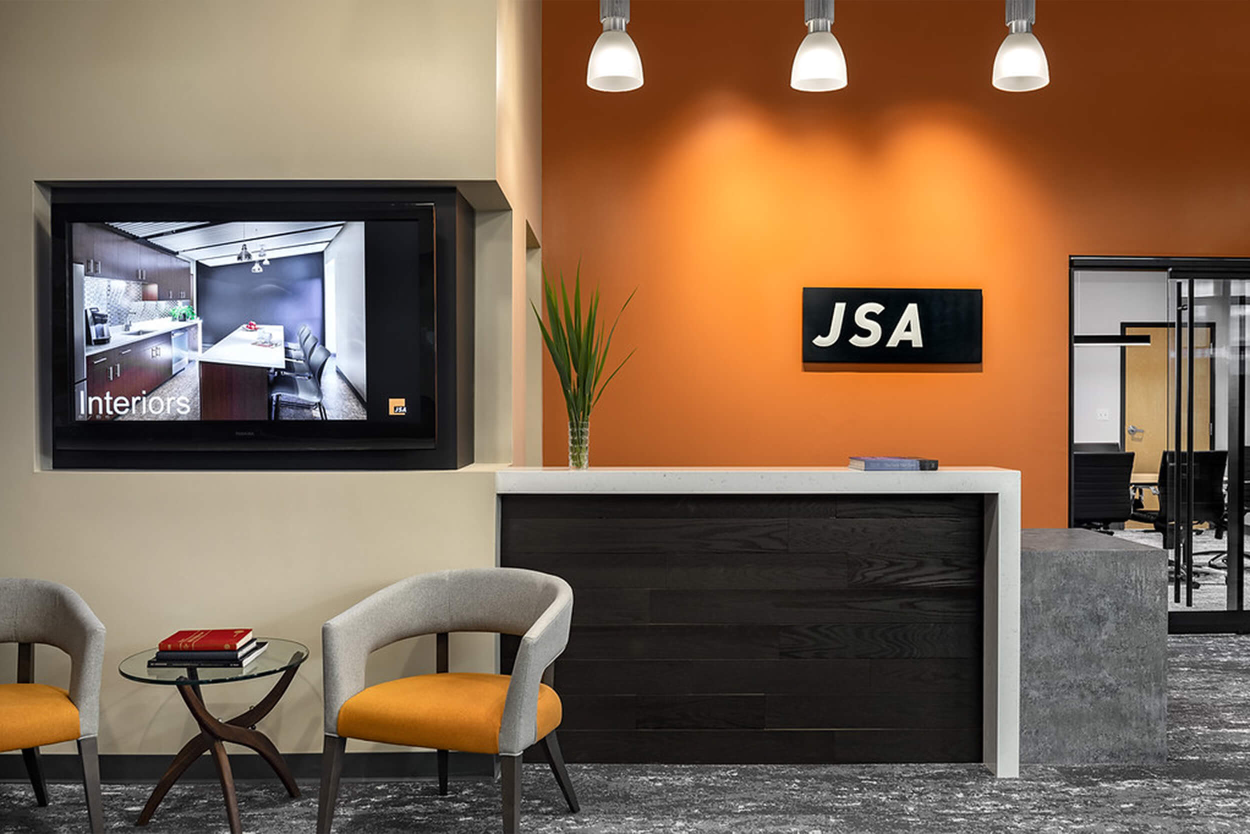 Photo of the interior entryway of JSA Inc's office. The photo features a dark wood paneled front reception area desk, with a bright orange wall behind it with a logo plaque of "JSA". To the left of the desk, there is a small seating area with two orange and grey chairs and an end table, and a video monitor on the wall is showing photos of interior design work.