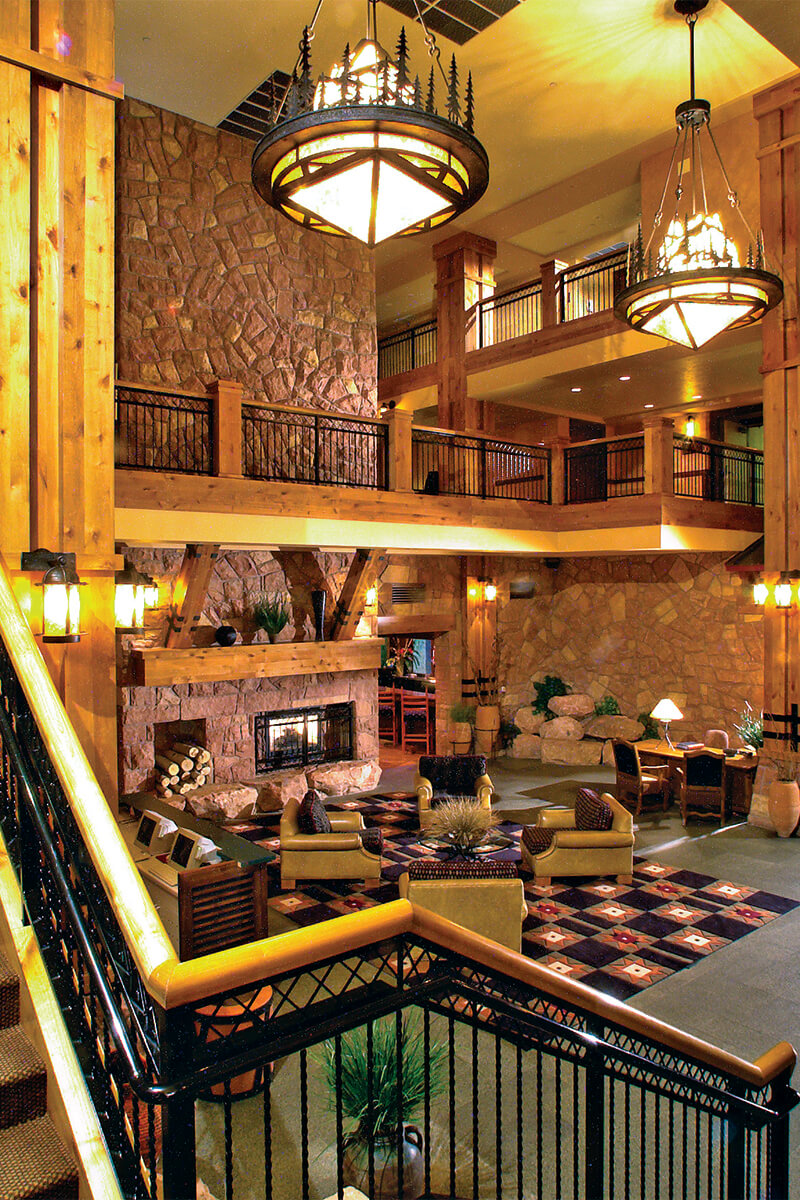 Interior view of a lobby staircase in a ski resort. Stone tile walls with wood trim create a warm and cozy atmosphere, with upholstered seating facing around a fireplace.