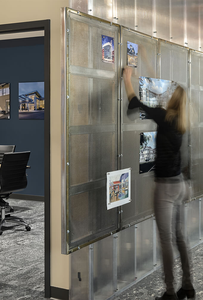 View of a woman (female JSA employee) in a dark shirt and grey jeans pinning up print-outs of architecture/interior design work onto a grey metal mesh board on an office wall.