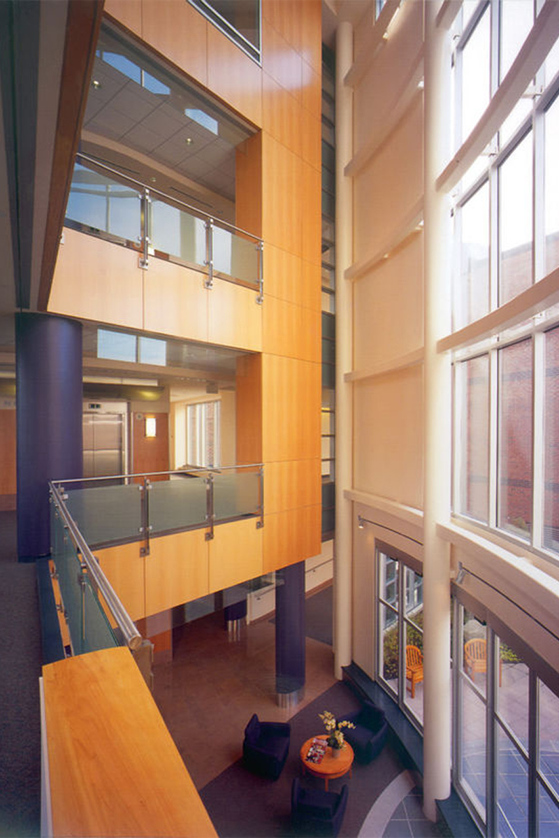 Interior view of lobby area atrium at a hospital facility. Wood paneling frames the floors above the ground floor, and large floor-to-ceiling windows are letting in lots of natural light.