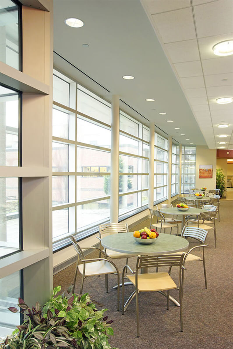 Interior view of a cafeteria at a hospital facility. Large floor to ceiling windows on the left let in lots of natural light, and small light blue tables with seating are shown.
