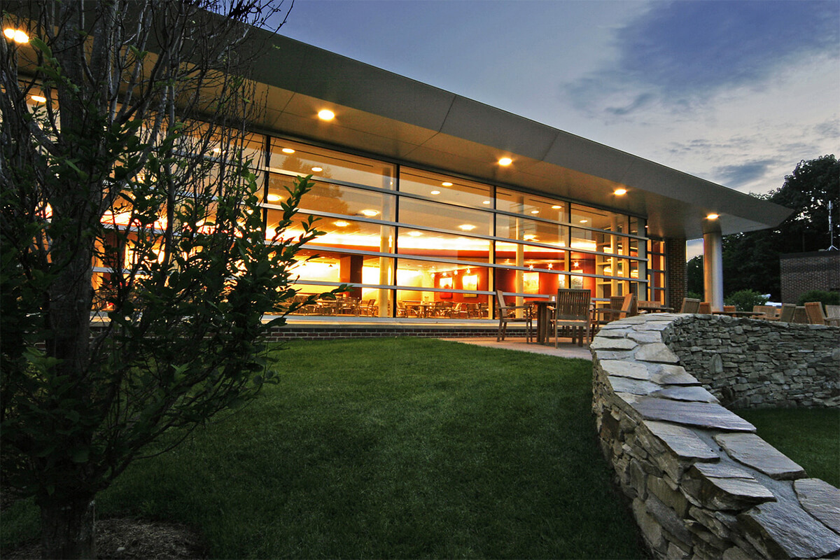 Exterior twilight photo of the cafeteria at a hospital facility. Lighting illuminates the building from within. A grassy area and low stone wall are outside the cafeteria which features large floor-to-ceiling windows.