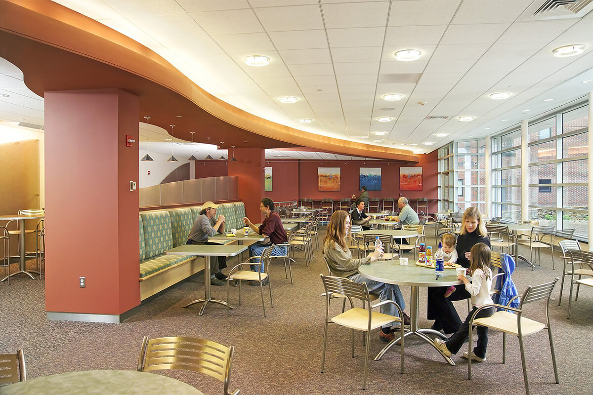 Interior view of a cafeteria at a hospital facility. A wavy light red center column wall features light blue and neutral colored dining banquettes. Several small tables and dining chairs are seen as well, and patrons are enjoying meals.