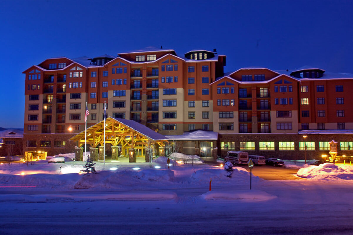 Nighttime exterior photo of a ski resort. Snow is seen in the foreground entryway of the building, which features a portico entrance. The multi-unit building has a brown and white facade.