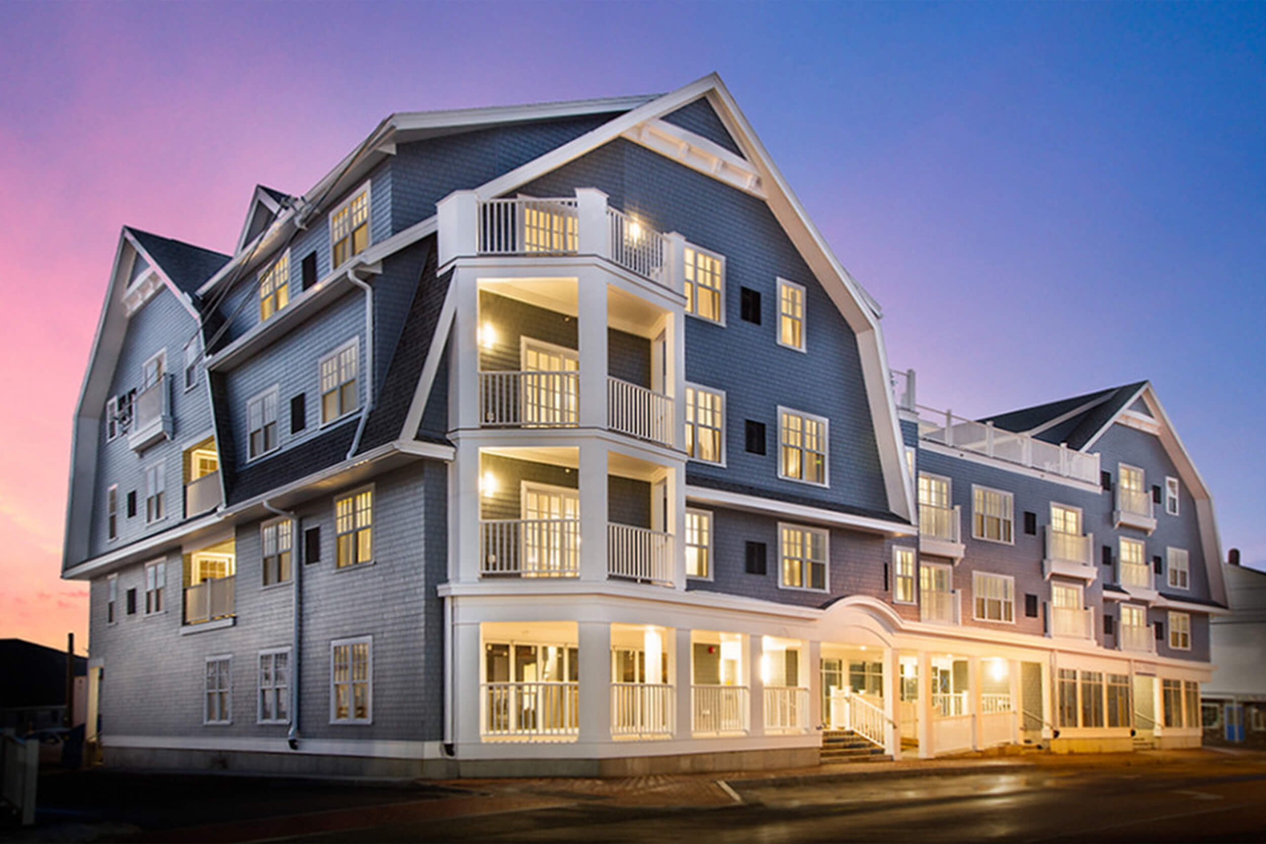 Exterior twilight photo of a beach club residence building. The four story building features blue siding with white trim, balconies, and an illuminated front porch area.