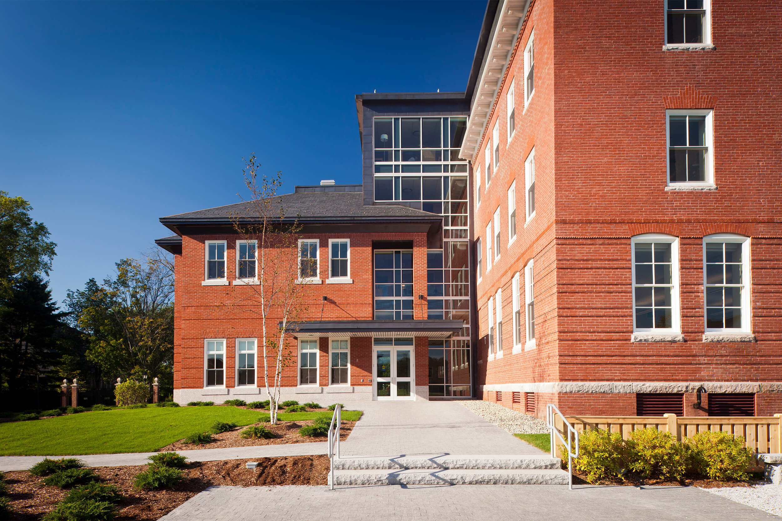 Exterior view of the entrance to Roger Williams Hall building at Bates College. A concrete walkway leads up to a small grass courtyard area by the entrance of the red brick building.
