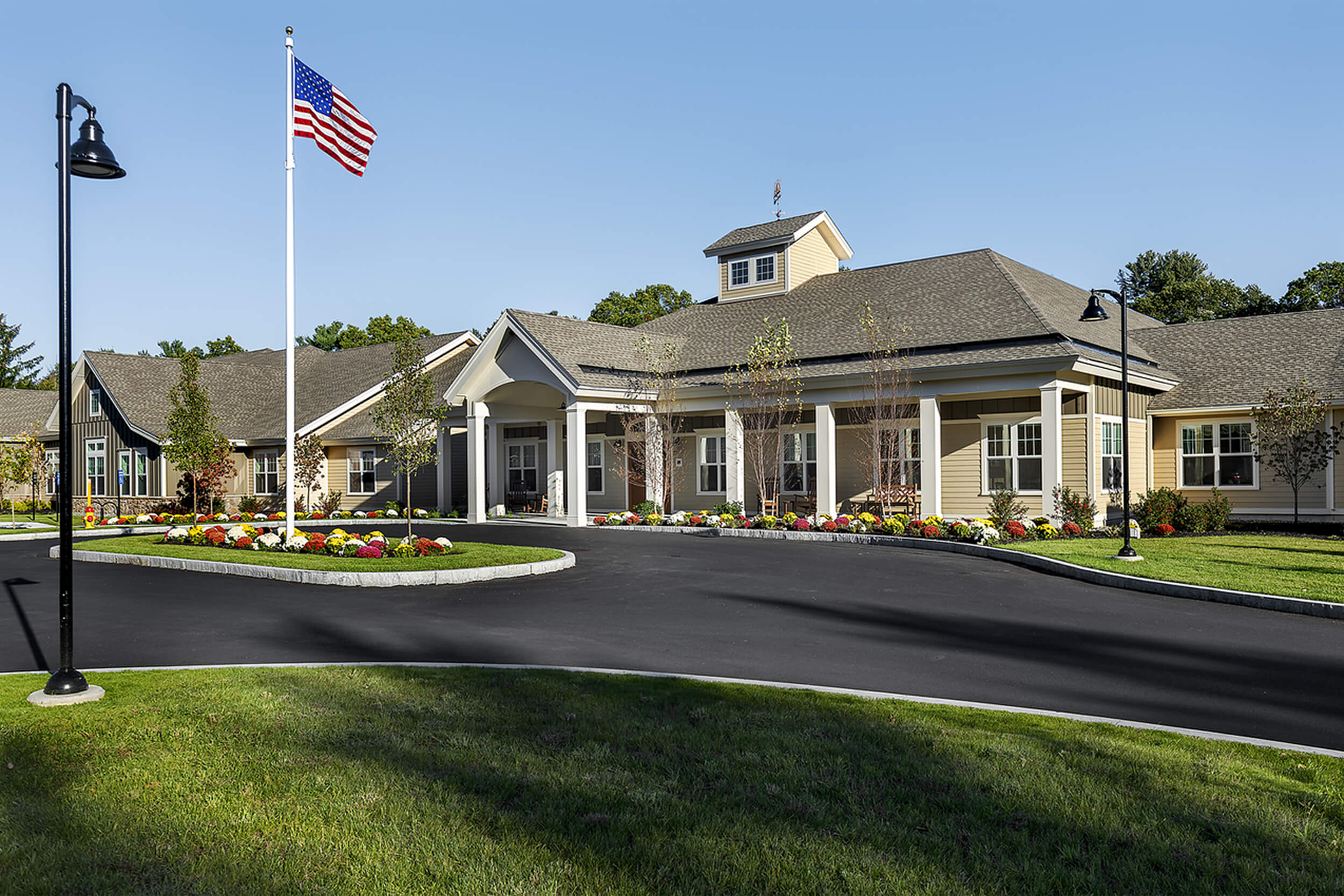 Exterior daytime view of the entry of an assisted living facility. The building is low and long, featuring neutral beige colored siding with white trim and a portico entry area. A drive-thru parking area is seen in front, with an American flag atop a flagpole in the middle grassy area.