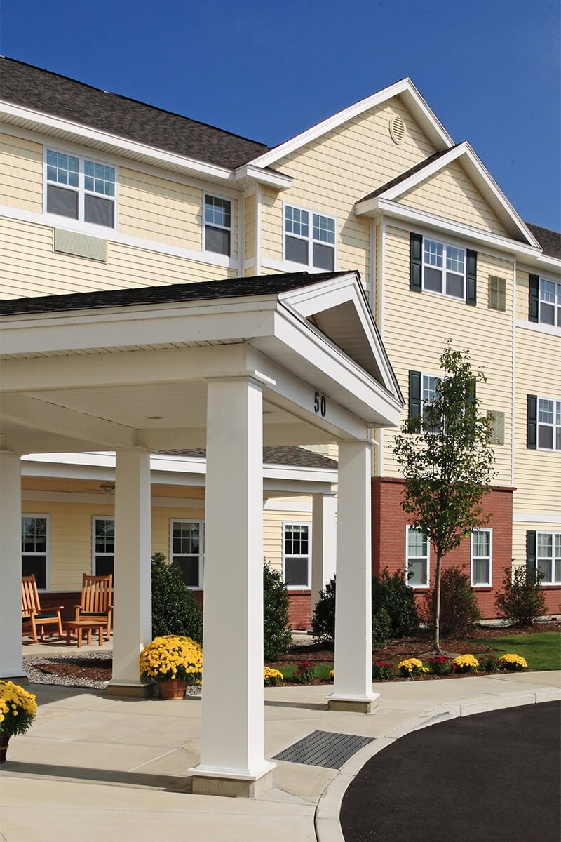 Exterior daytime view of a portico entryway to a senior living facility. The building has light yellow beige siding with red brick accents at the bottom and white trim.