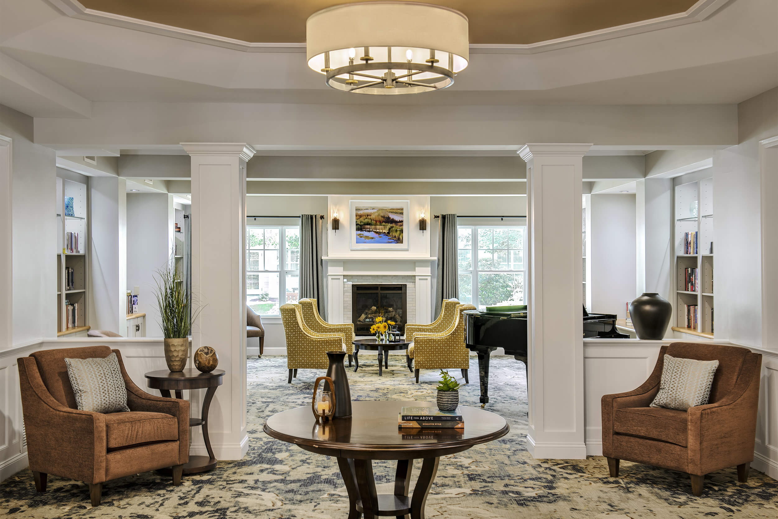 Interior view of a lobby at a senior living facility. An open area features several upholstered lounge chairs, a circular table in the middle with decorative centerpieces, and in the background a fireplace can be seen with yellow upholstered chairs around it.
