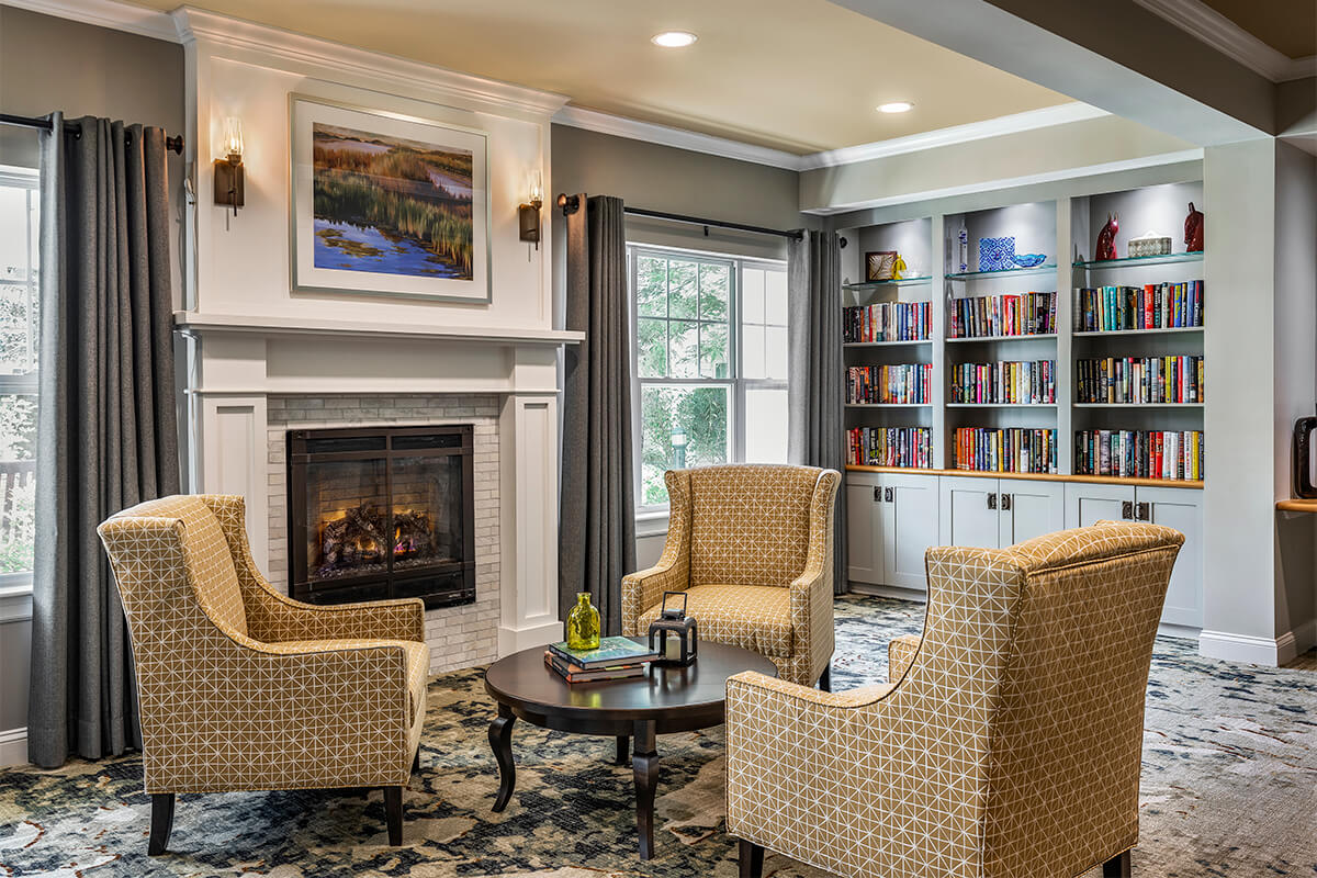 Interior view of a seating area with small library at a senior living facility. Several beige patterned upholstered chairs surround a low circular wood coffee table net to a fireplace. Large windows let natural light spill in, highlighting a built-in shelving unit in the background that houses many colorful books.