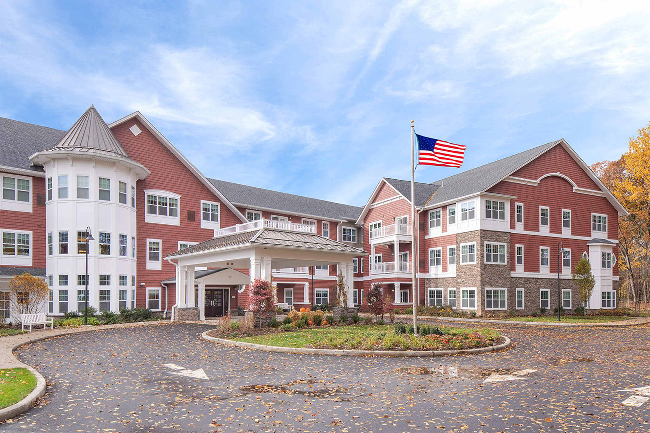 Exterior daytime view of a senior living facility entrance. The buildings feature red shingle siding with white trim. A roundabout driveway is in front of the entrance, with an American flag atop a flagpole in the center grassy area.