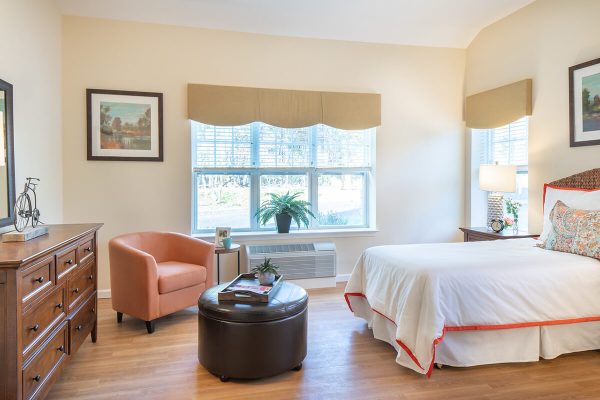 Interior view of a resident room at a senior living facility. The room is bright and airy with natural light coming from a widow on the far wall, neutral colored walls, and light wood flooring. Bedroom furniture including a bed, dresser, small table, and upholstered chair can be seen.