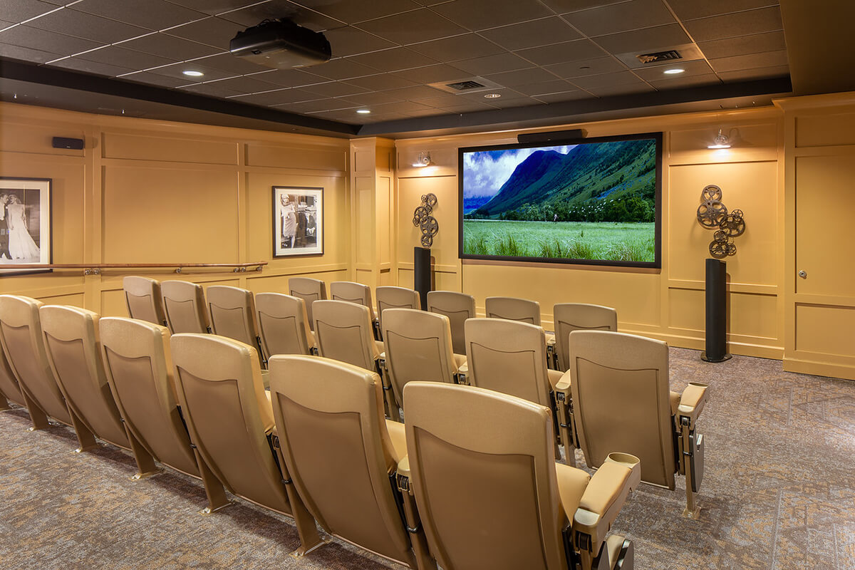 Interior view of a media room at a senior living facility. Several rows of tan leather theater chairs are seen facing a large TV monitor mounted to a beige colored wall.