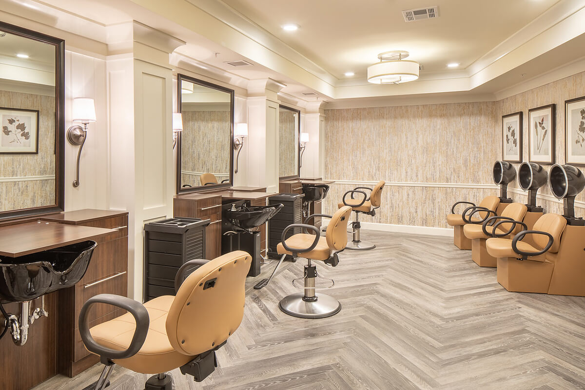 Interior view of a salon at a senior living facility. The floor is a herringbone grey wood pattern, and several hairdressing stations and sink washing stations with light tan leather seats are seen.