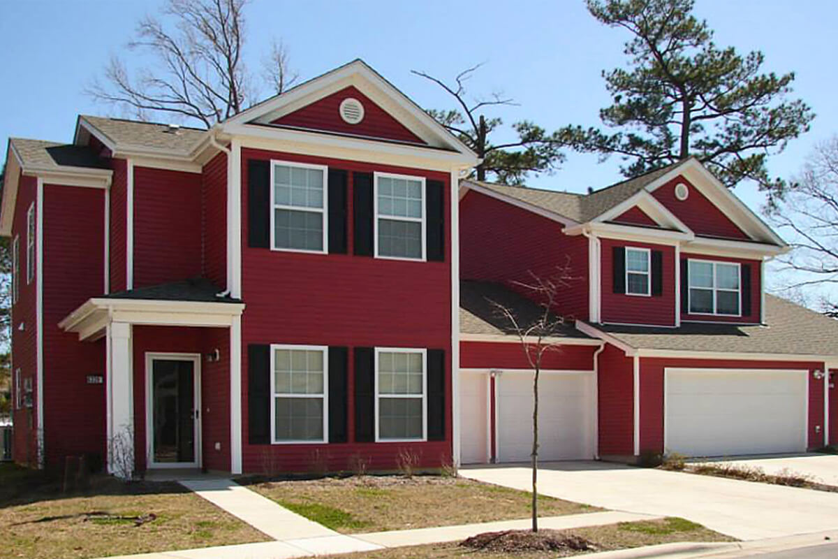An angled exterior view of a multi-family house at Camp Lejeune. The house features red siding with white trim, large windows, black shutters, two white garage doors in the middle and entryways on the left and right side.
