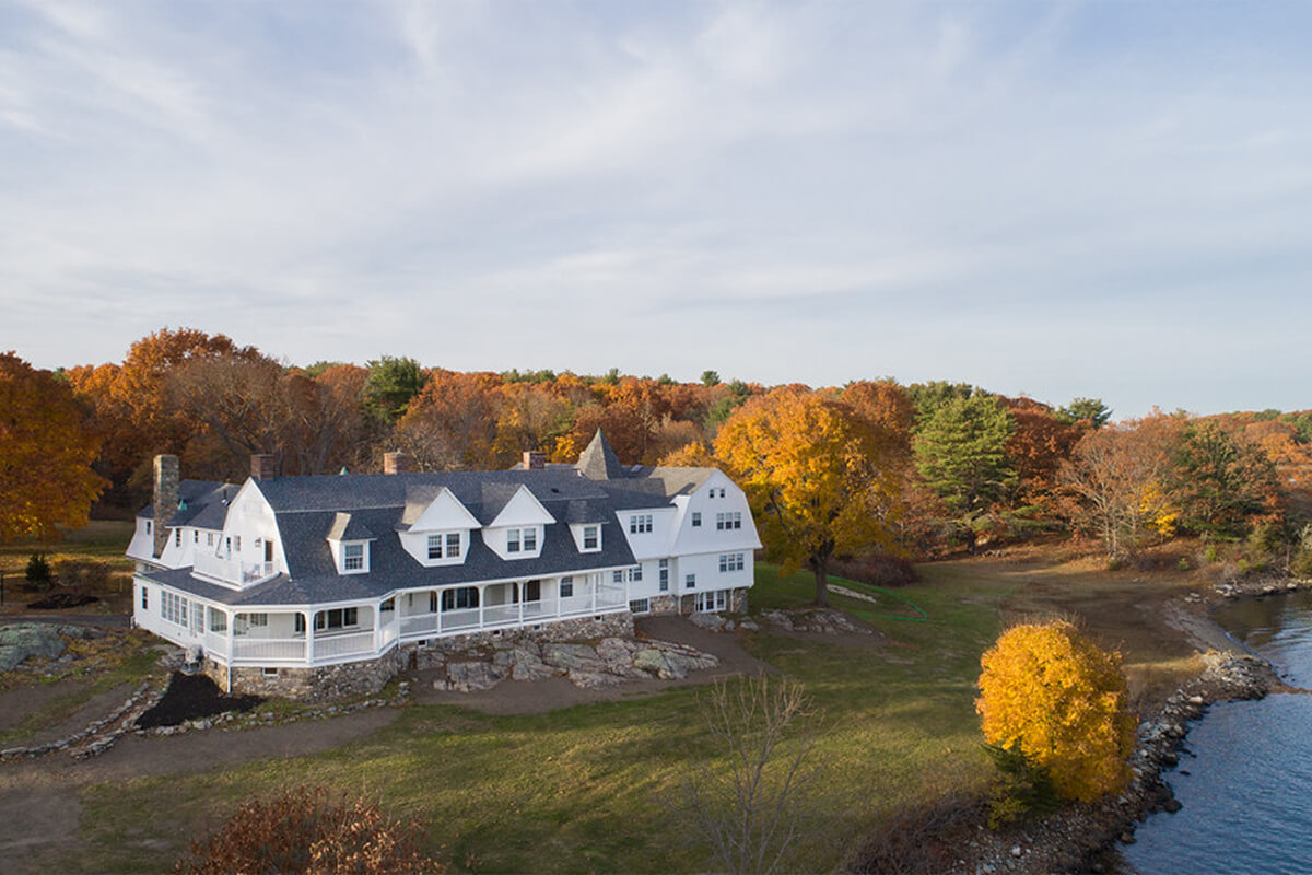 Exterior daytime drone photo of a beautiful white historical building with a large porch and pitched roof. Behind the building are beautiful fall-colored trees, and a body of water sits to the right of the building.