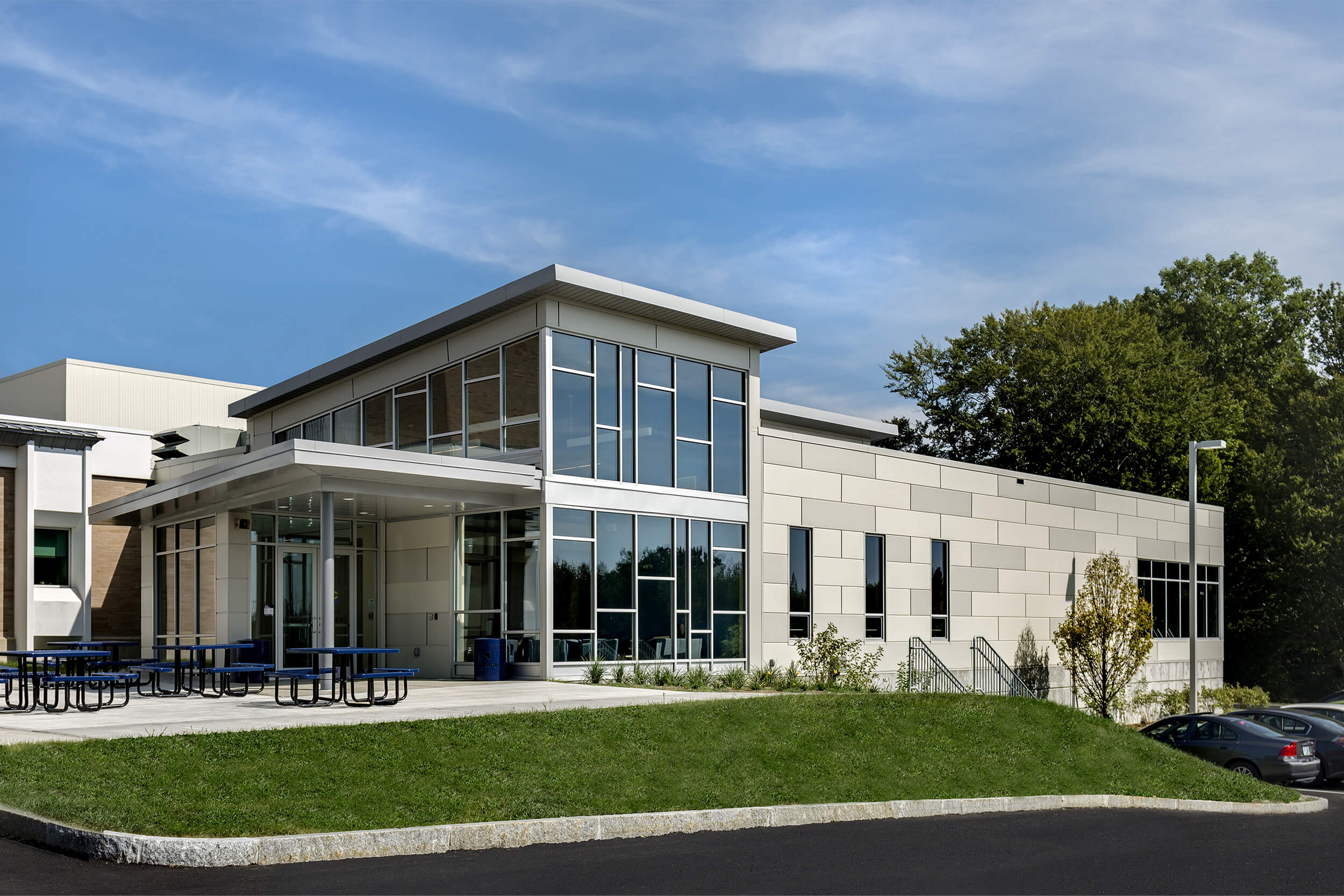 An exterior view of the student success center at Great Bay Community College. The building is a low grey stone building with two floors, open glass windows/walls on the exterior entrance, and a small courtyard with blue metal picnic tables.