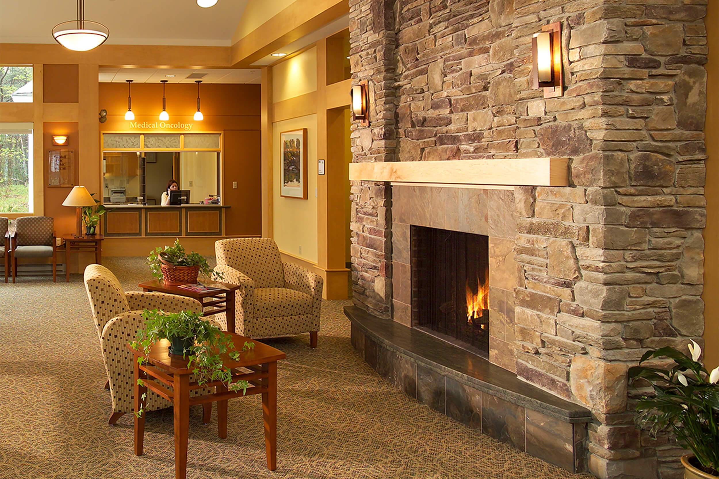 Interior photo of a waiting area at an oncology treatment center. A stone fireplace is seen in the foreground with two comfortable chairs and end tables facing it, and further in the background a wood paneled reception desk area can be seen.