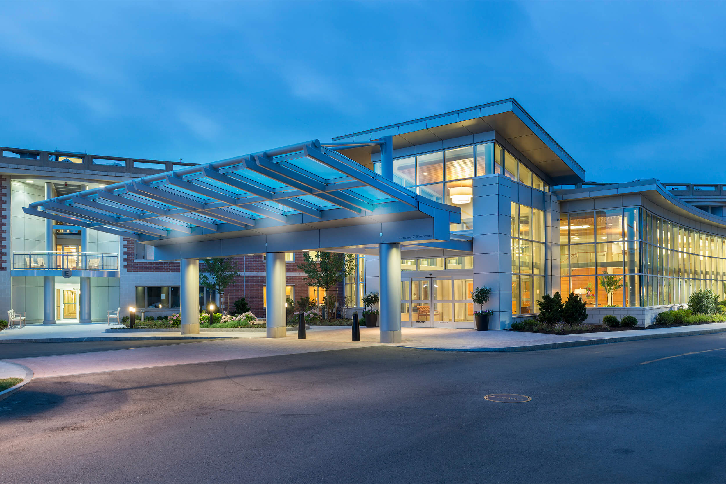 Exterior view of the entry of a senior living facility building. The photo is taken at dusk, with lighting inside the building illuminating it. The entryway features large floor-to-ceiling glass walls/windows.